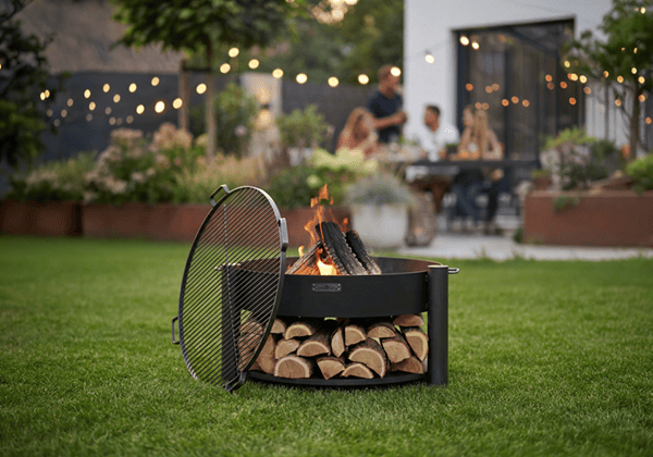 Brasero de jardin avec grille et bois stocké, posé sur l’herbe, ambiance potager et éclairage festif en arrière-plan.