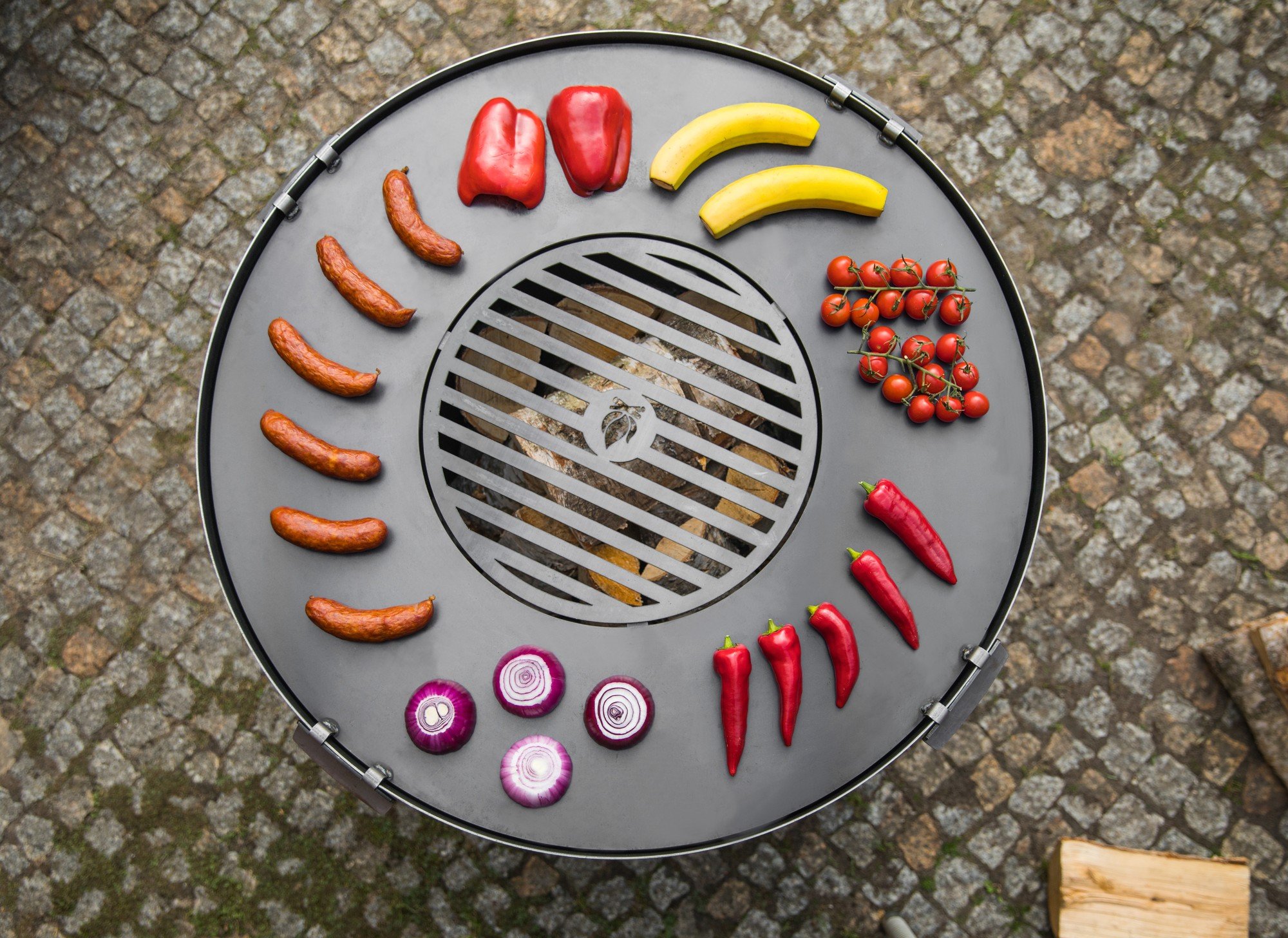 Planche de barbecue ronde vue d’en haut avec des légumes et saucisses disposés autour du gril, sur pavés extérieurs.
