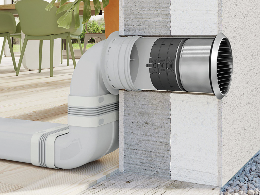 Système de ventilation pour hotte de cuisine avec conduit flexible en PVC gris et boîtier noir, installé dans un mur, près d’un espace intérieur avec mobilier vert.